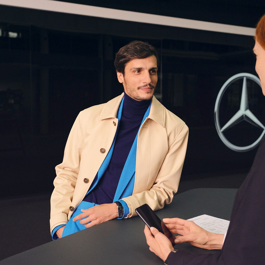 ‎ A customer leaning at the counter turns expectantly to face a Mercedes-Benz employee.
