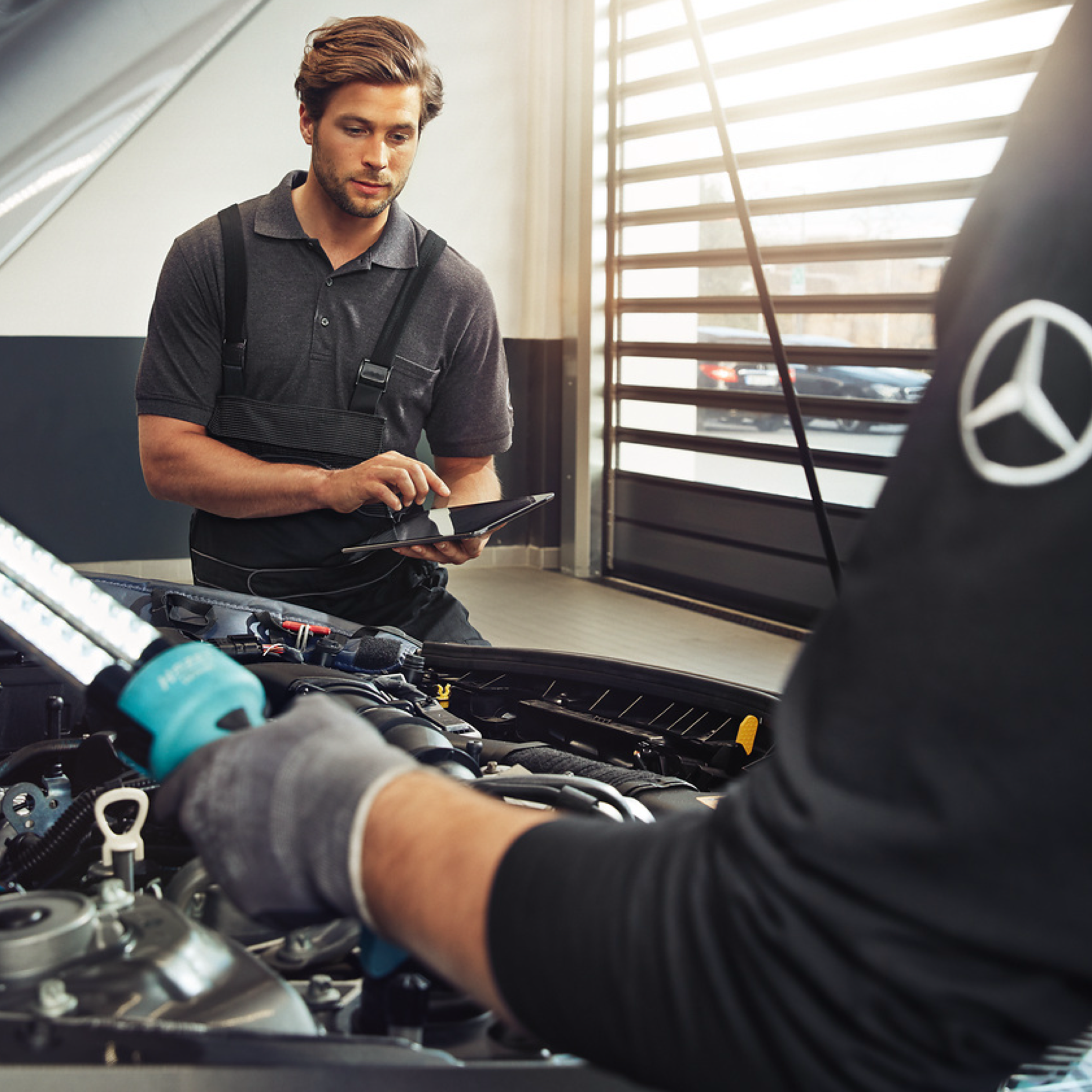 GenuineParts Two Mercedes-Benz technicians check the engine compartment of a vehicle.