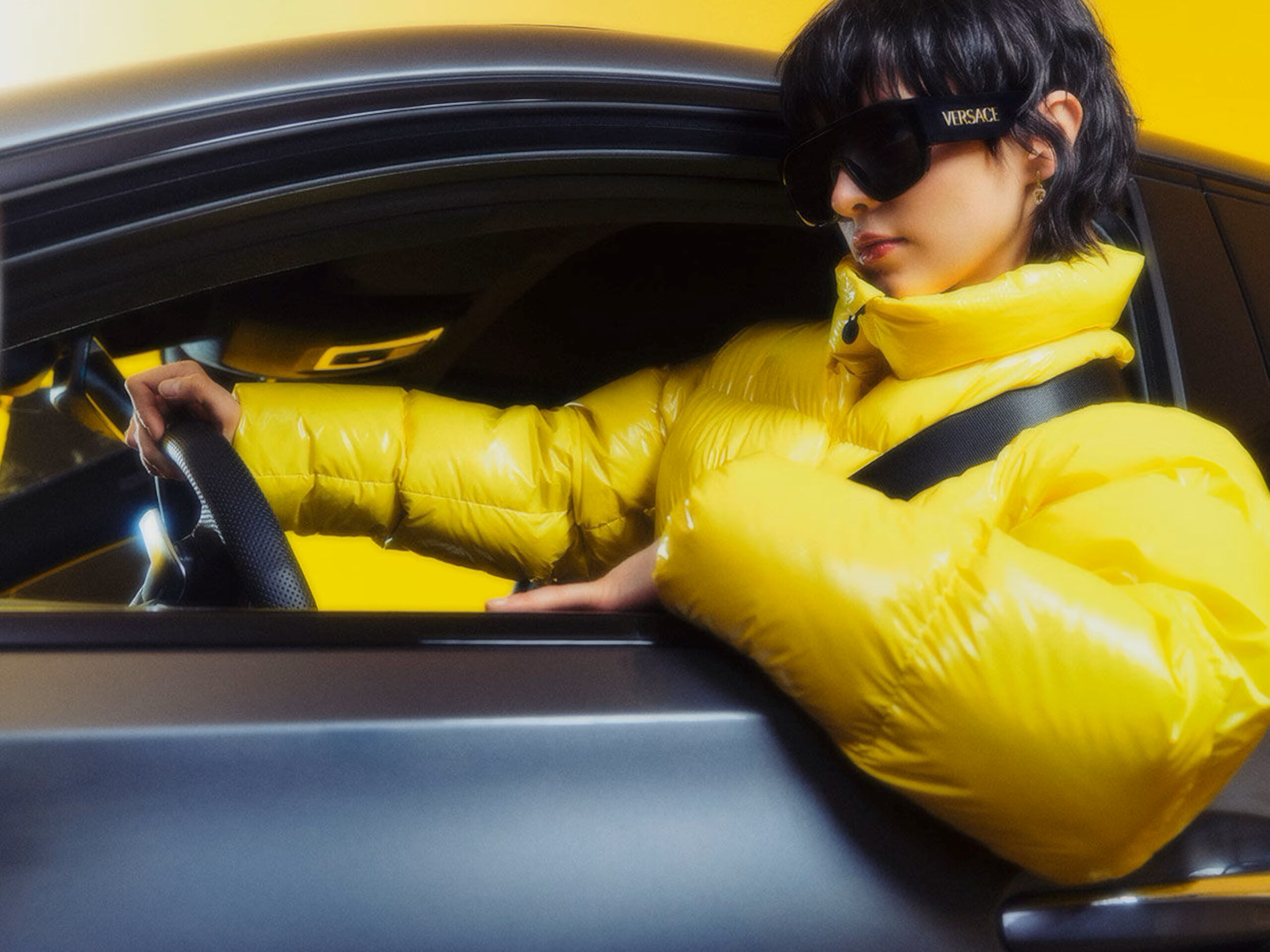 A woman in a white gown and a man standing behind in a sleeveless top and white pants with a sculpture behind featuring the Mercedes-Benz logo